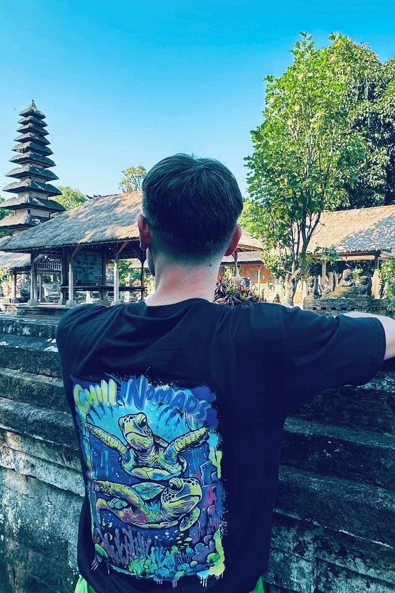 Person wearing a black spicy lotus t-shirt with a colorful turtle design, standing in front of traditional balinese architecture.