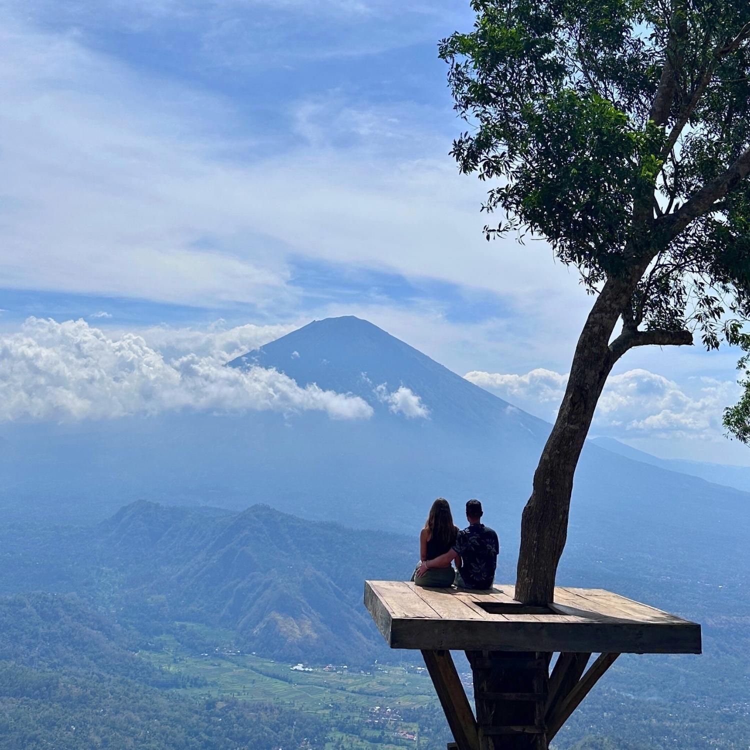 Two people sitting on a wooden platform overlooking Mount Agung volcano at lahangan sweet viewpoint in Bali, Indonesia with a clear blue sky.