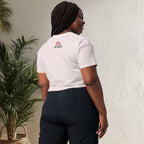 Person wearing a white crop top with a logo on the back, standing against a light-colored wall with a plant to the left.