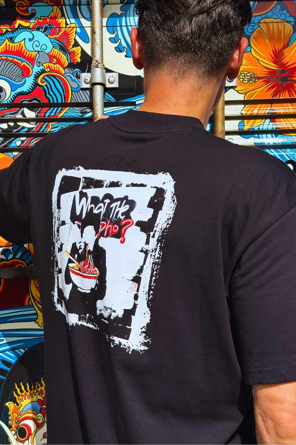 Male model wearing a black Spicy Lotus t-shirt with a pho noodle graphic design on the back, standing in front of a colorful mural.