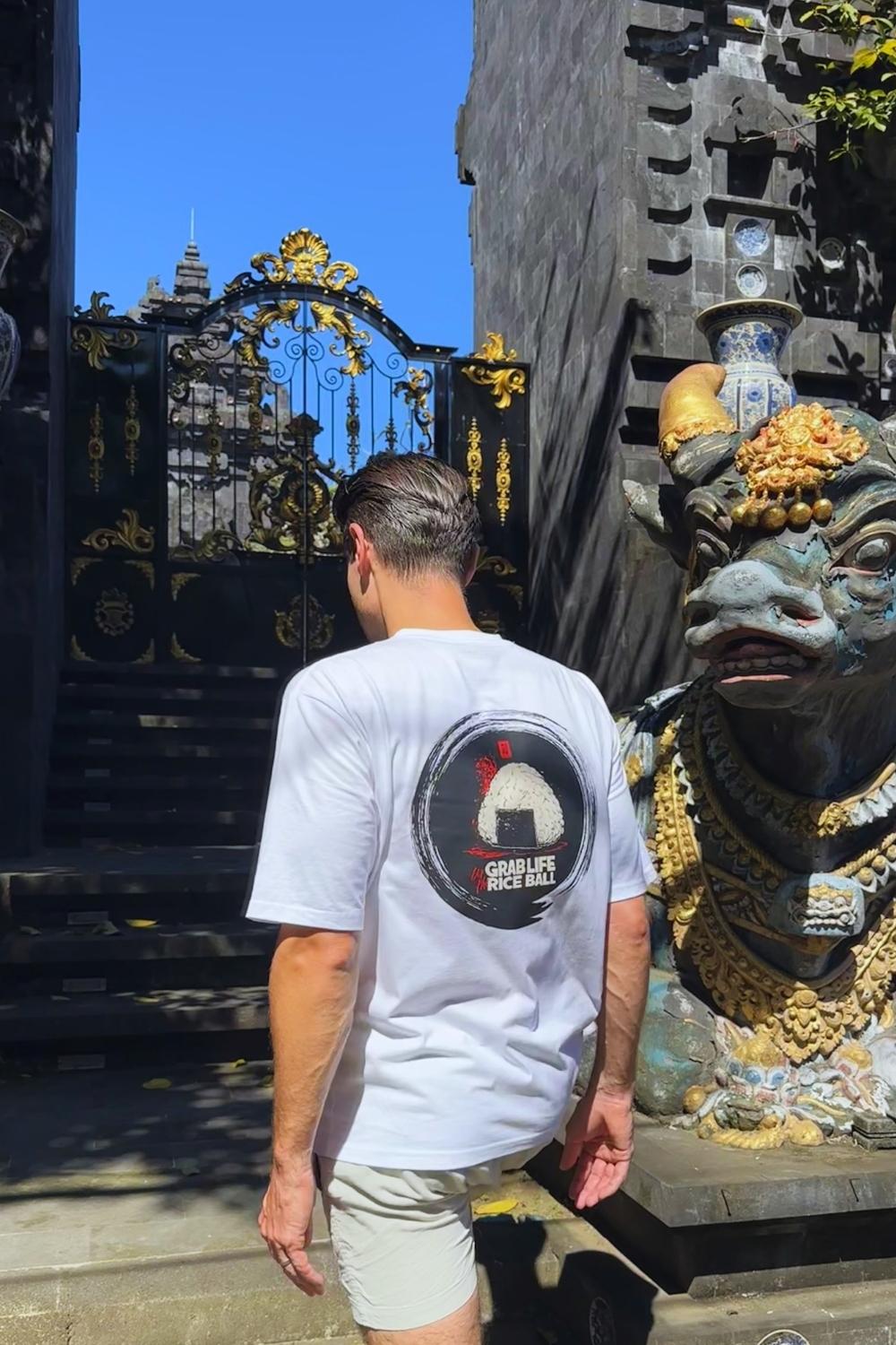 Male model in a white t-shirt with a logo standing in front of a large, ornate gate and statue.