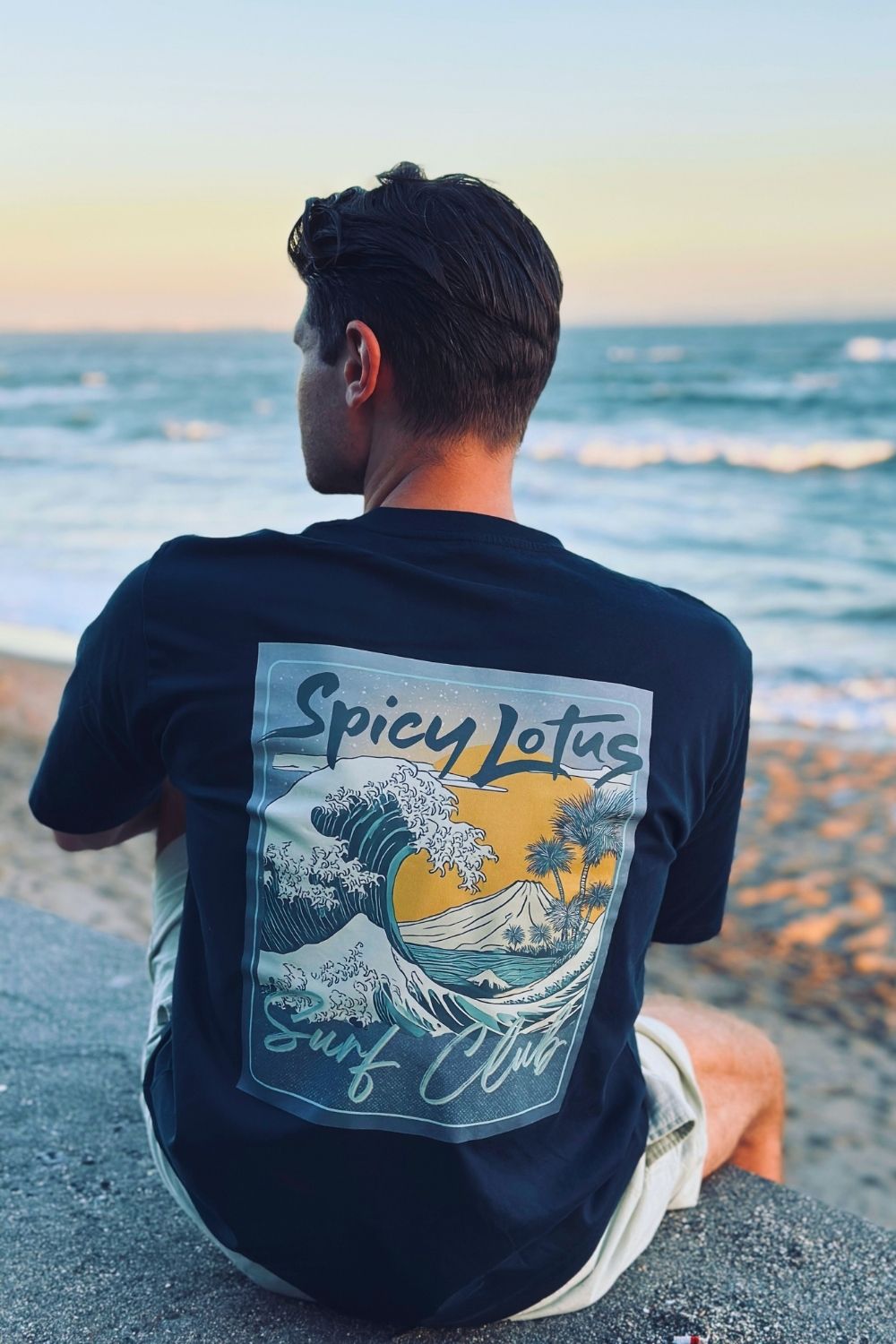 Person wearing a 'Spicy Lotus Surf Club' t-shirt sitting on a beach.