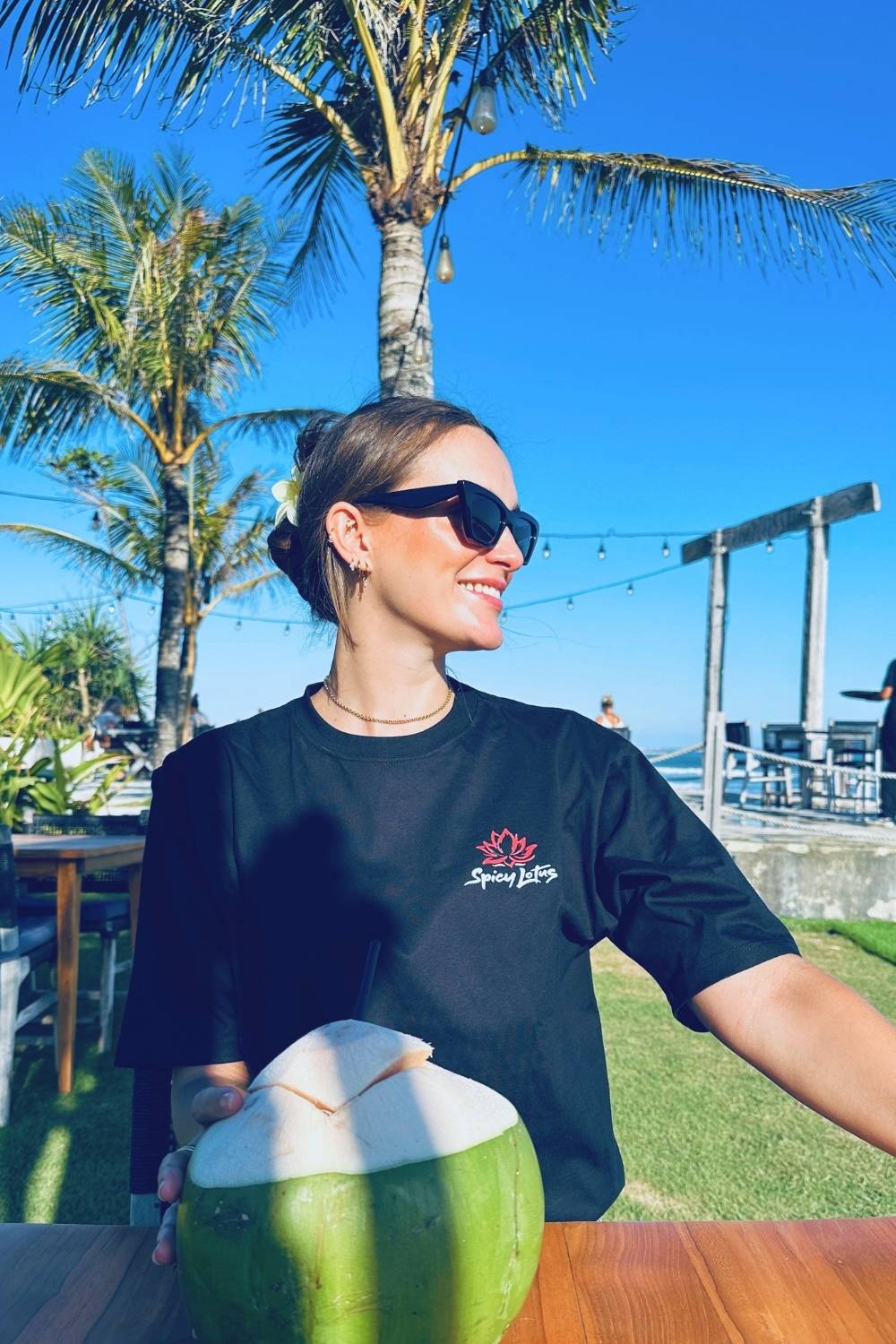 Woman holding a green coconut outdoors with palm trees and clear blue sky in the background wearing a spicy lotus t-shirt.