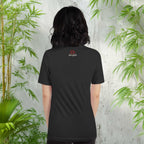 Person wearing a black t-shirt with a logo in front of a green plant and light gray wall.