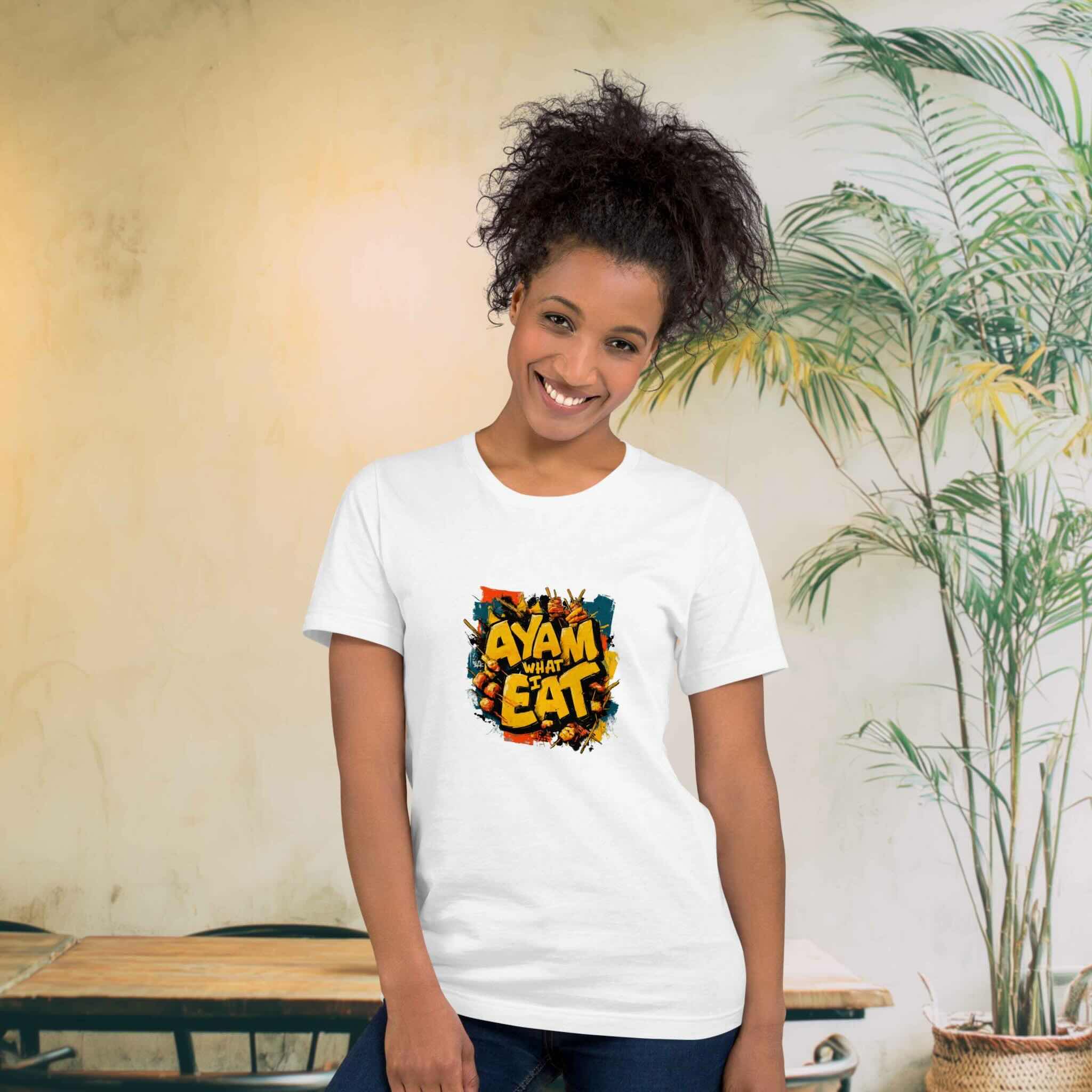 Woman wearing a white t-shirt with colorful text design, standing indoors with a plant in the background.