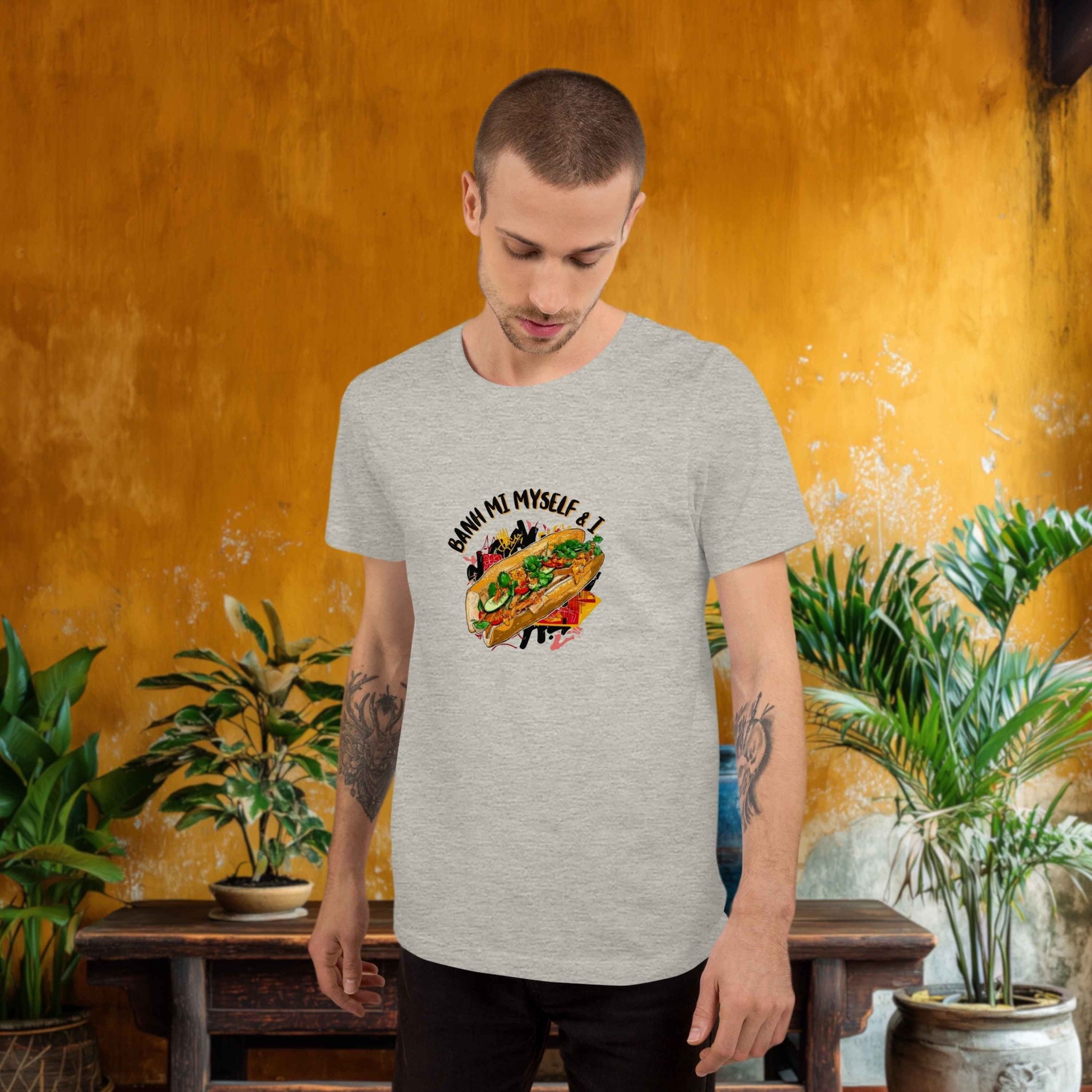 Man wearing a gray t-shirt with a graphic design of a Banh MI baguette in front of a textured orange wall with plants.