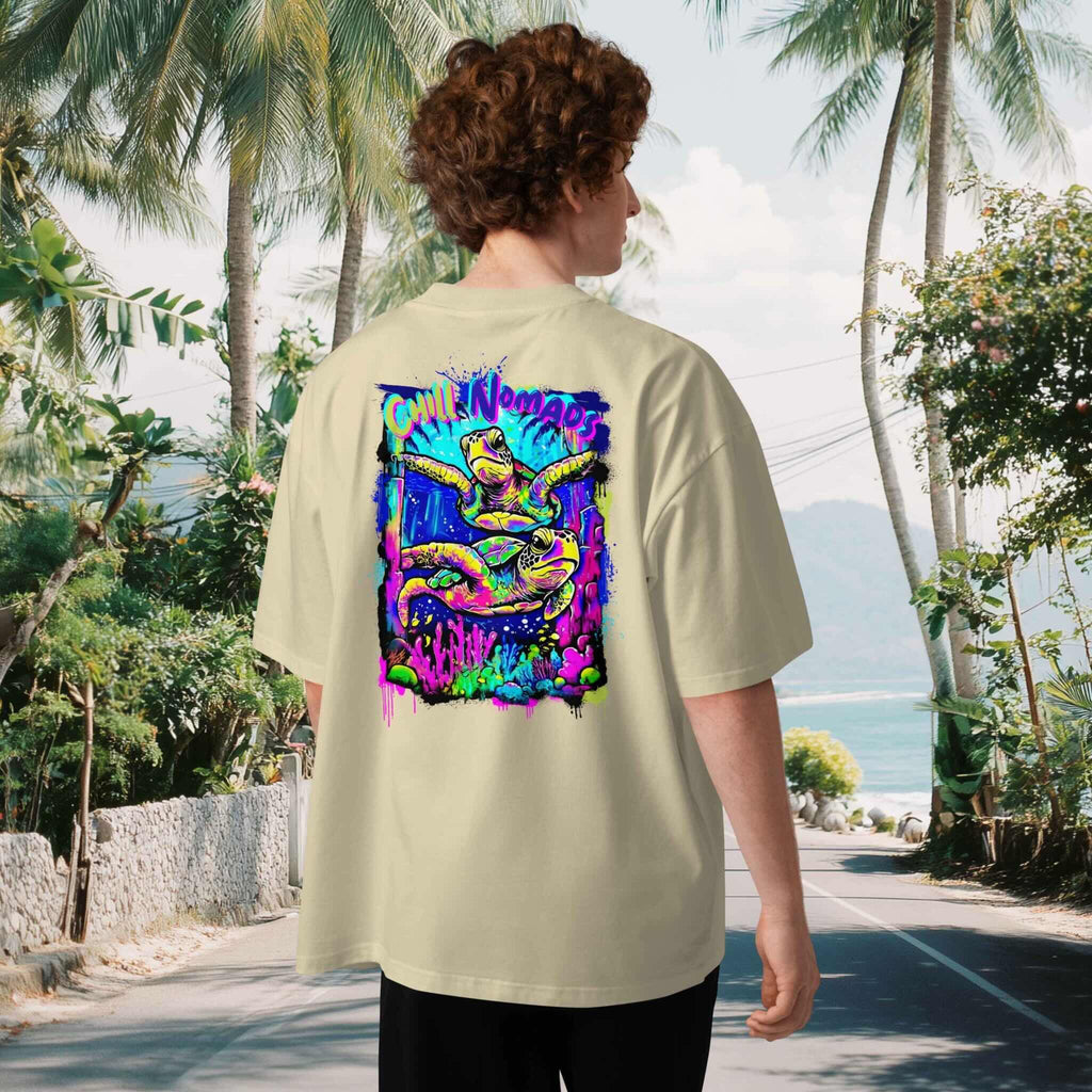 Man wearing a beigeoversized t-shirt with a colorful graphic of two turtles on the back, standing in a tropical setting with palm trees and a beach.