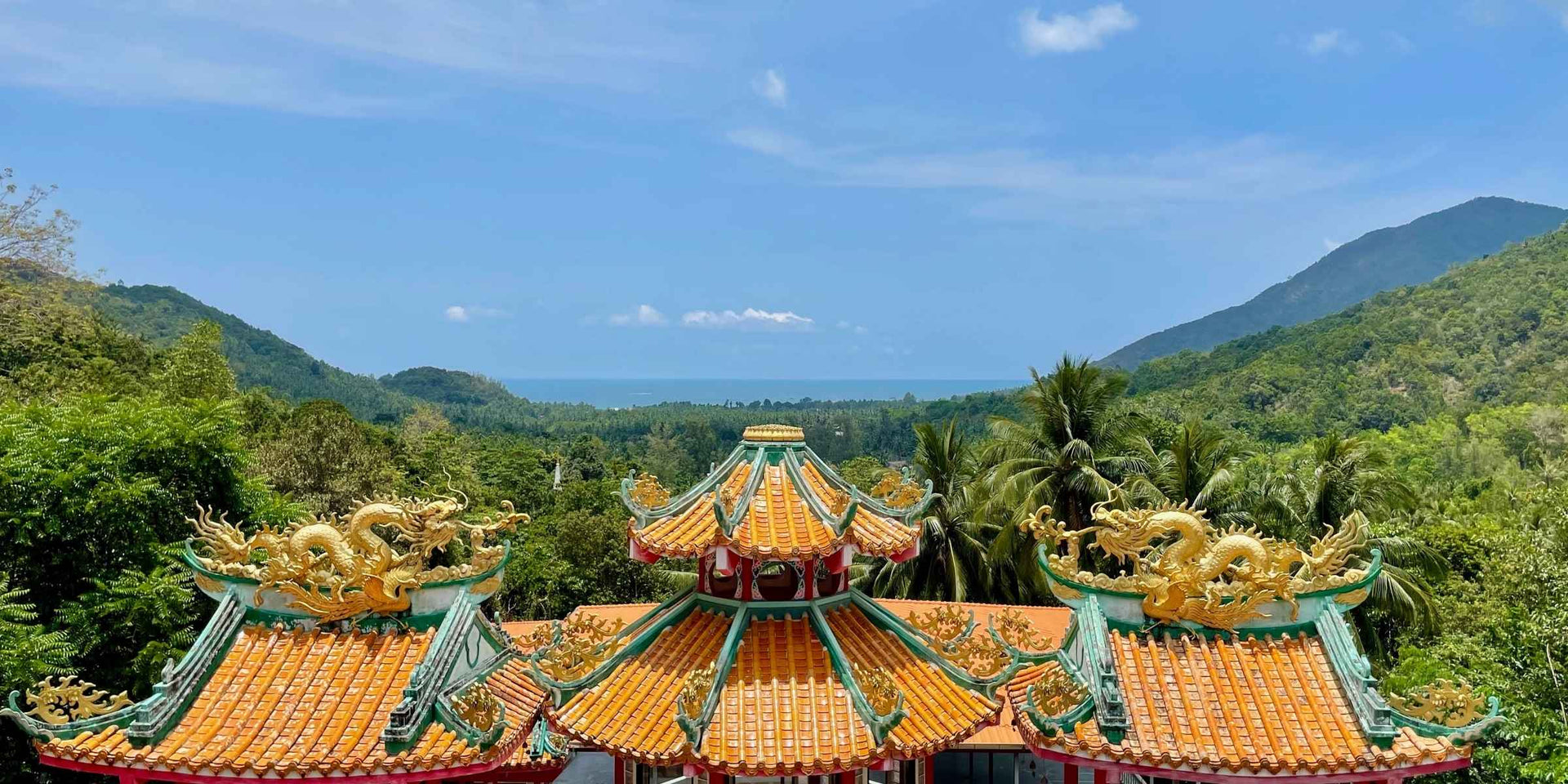 Traditional Chinese temple with golden dragons on the roof surrounded by green mountains, trees and the ocean in the background in Koh Phangan, Thailand