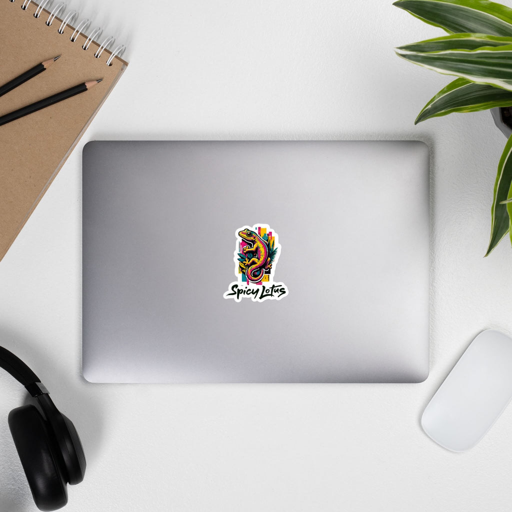 Laptop with a 'Spicy Lotus' sticker with a colorful gecko graphic on a laptop on white surface with office items.