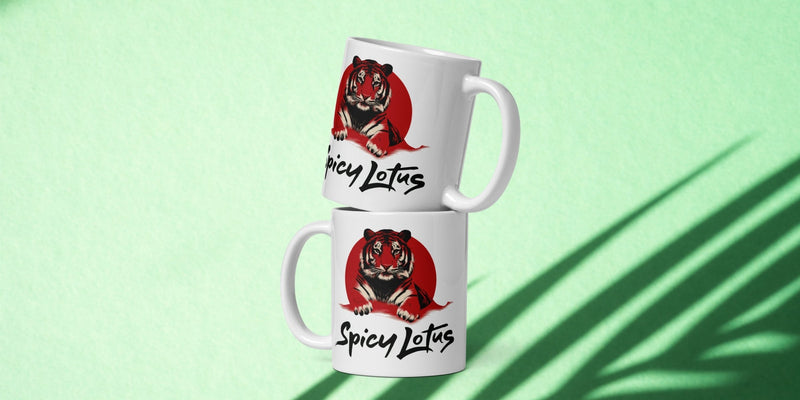 Two white mugs with a tiger design and 'Spicy Lotus' logo on a light green background wit tropical shadows.