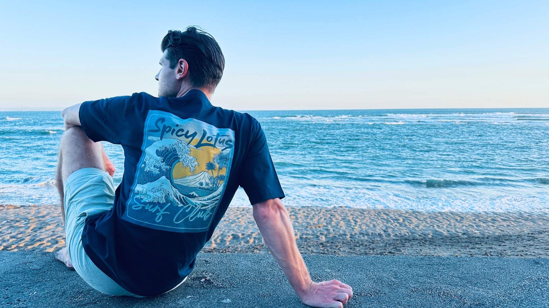 A man sits on a sandy beach facing the ocean, wearing a black graphic t-shirt with a large 'Spicy Lotus Surf Club' print on the back.