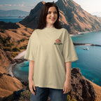 Woman wearing a beige oversized organic t-shirt with a lotus flower logo in front of a scenic mountain and ocean backdrop at Komodo Island viewpoint, Indonesia.