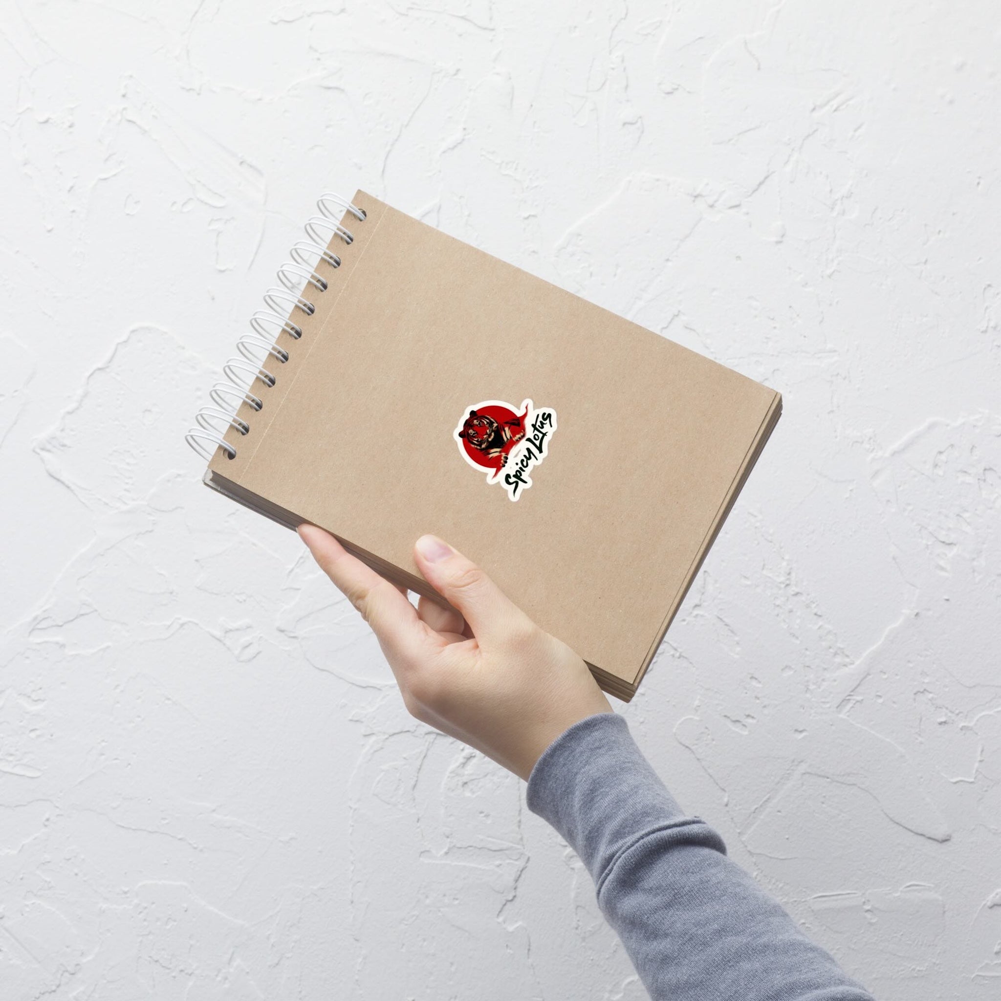 Hand holding a brown notebook with a red and white tiger sticker on a light gray background