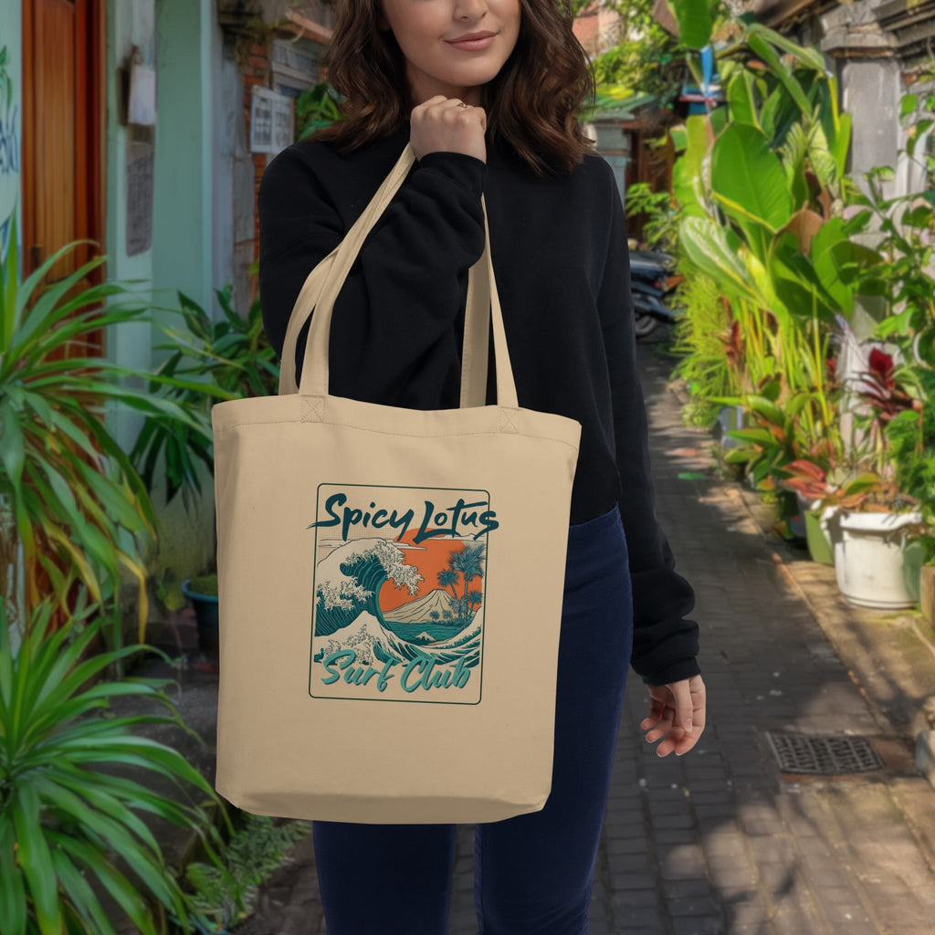 Woman holding a beige tote bag with 'Spicy Lotus Surf Club' design in a garden setting
