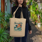 Woman holding a beige tote bag with 'Spicy Lotus Surf Club' design in a garden setting