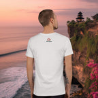 Man wearing a white t-shirt with a logo, standing on a cliff in Uluwatu, Bali overlooking the ocean at sunset.