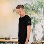 Man wearing a black t-shirt with a logo in a casual setting with plants and tables.