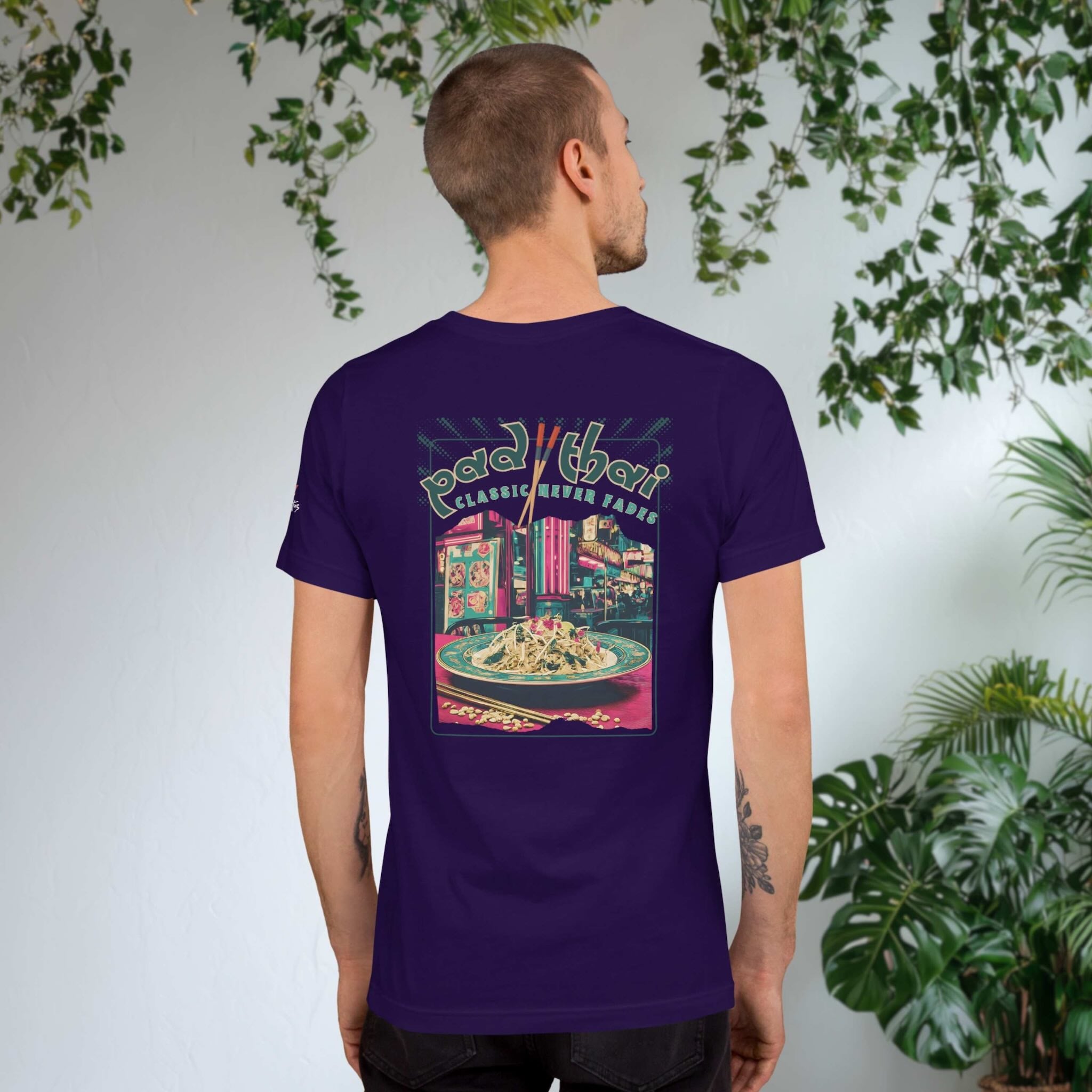 Man wearing a purple t-shirt with a colorful Pad Thai graphic on the back, standing against a light gray wall with greenery.