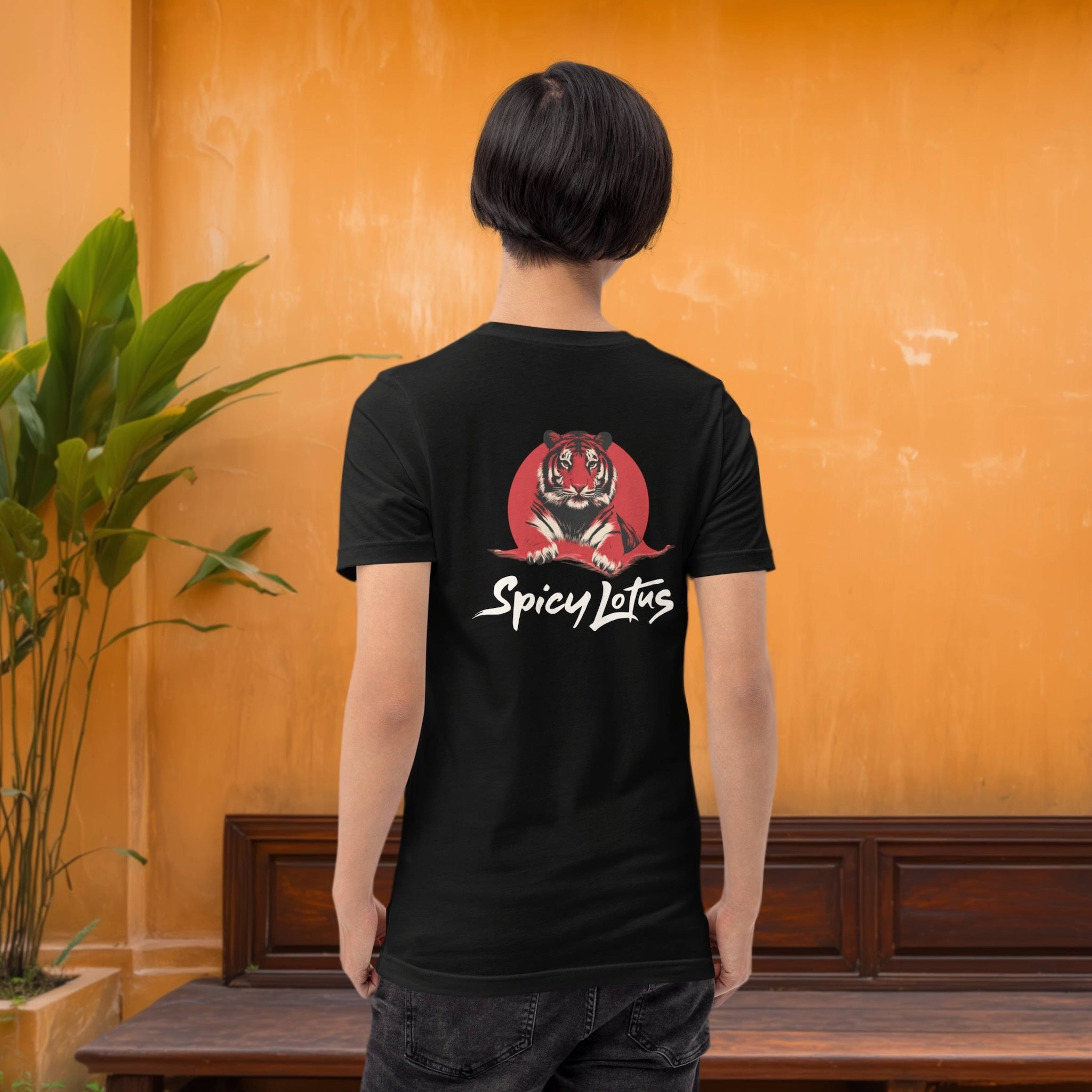 Person wearing a black t-shirt with 'Spicy Lotus' tiger design, standing against an orange wall.