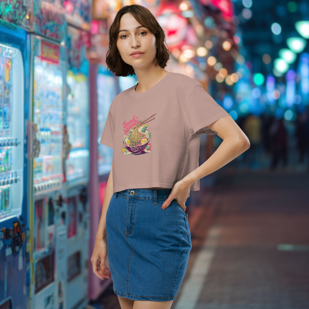 Woman wearing a pink crop top with a colorful graphic design of japanese ramen with the pun 'Send Noods'  in front of a neon-lit arcade.
