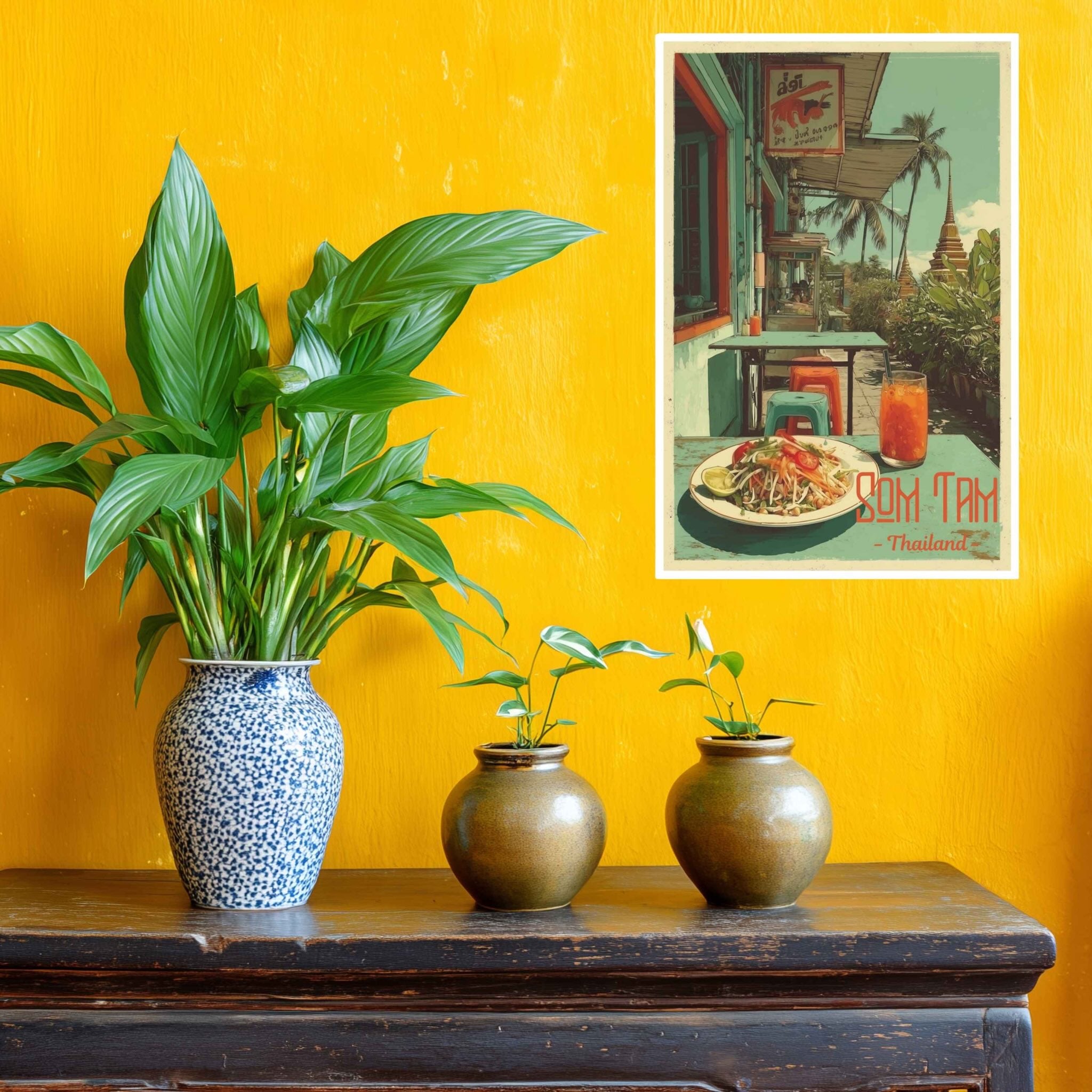 Three potted plants on a wooden table with a yellow wall and a framed picture of a tropical scene.