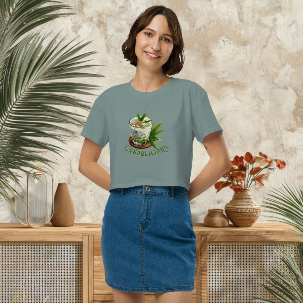 Woman wearing a light blue t-shirt with a graphic design of cendol dessert and text 'Cendolicious' standing in a room with plants and decor.