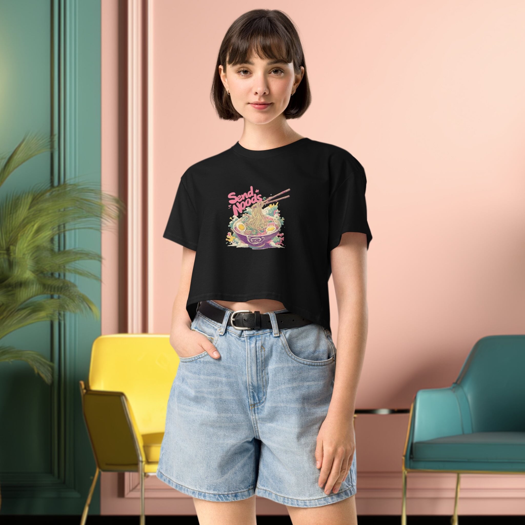 Person wearing a black crop top with colorful graphic design  of japanese ramen and denim shorts in a modern interior setting.