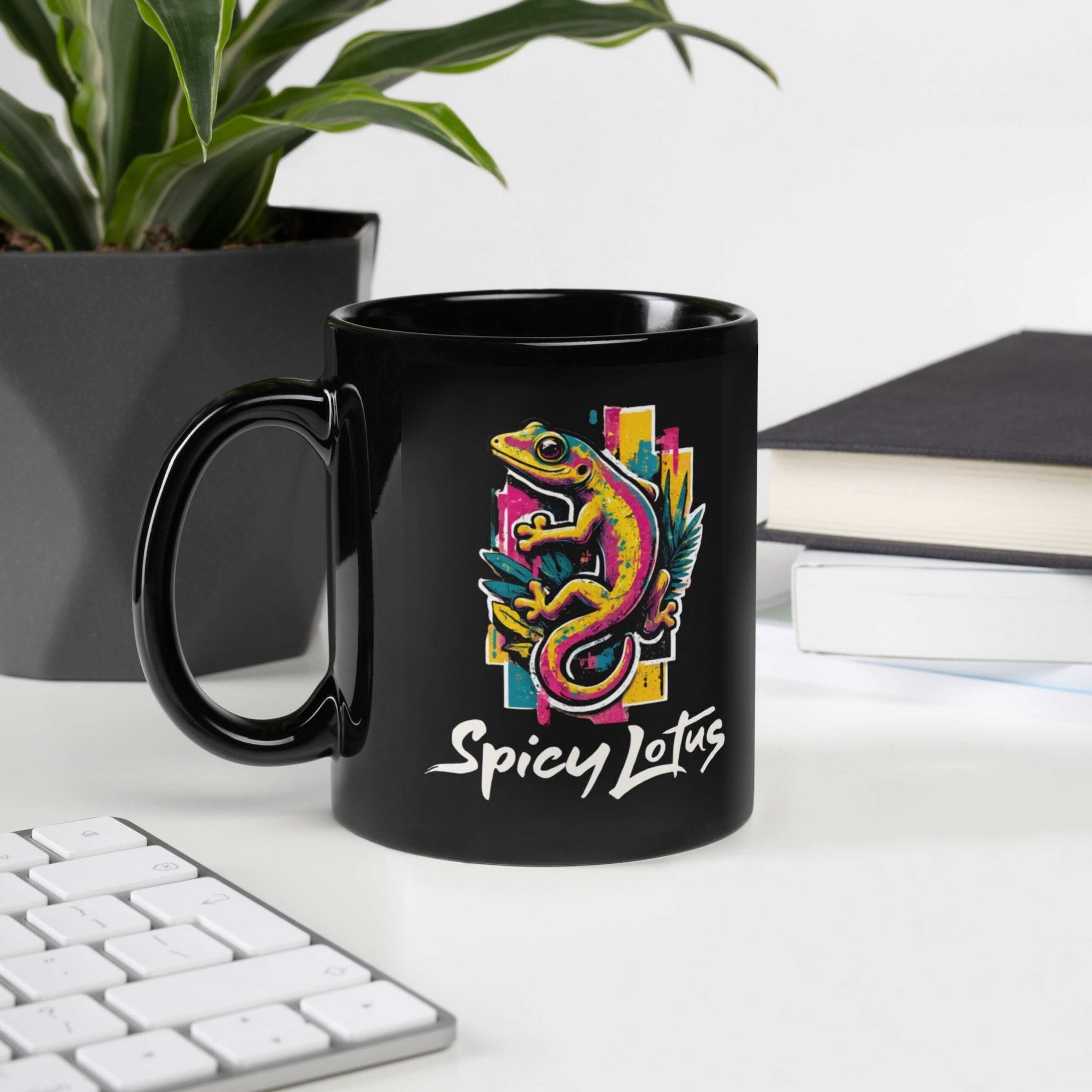 Black mug with colorful gecko design and 'Spicy Lotus' text on a desk with books and plant.