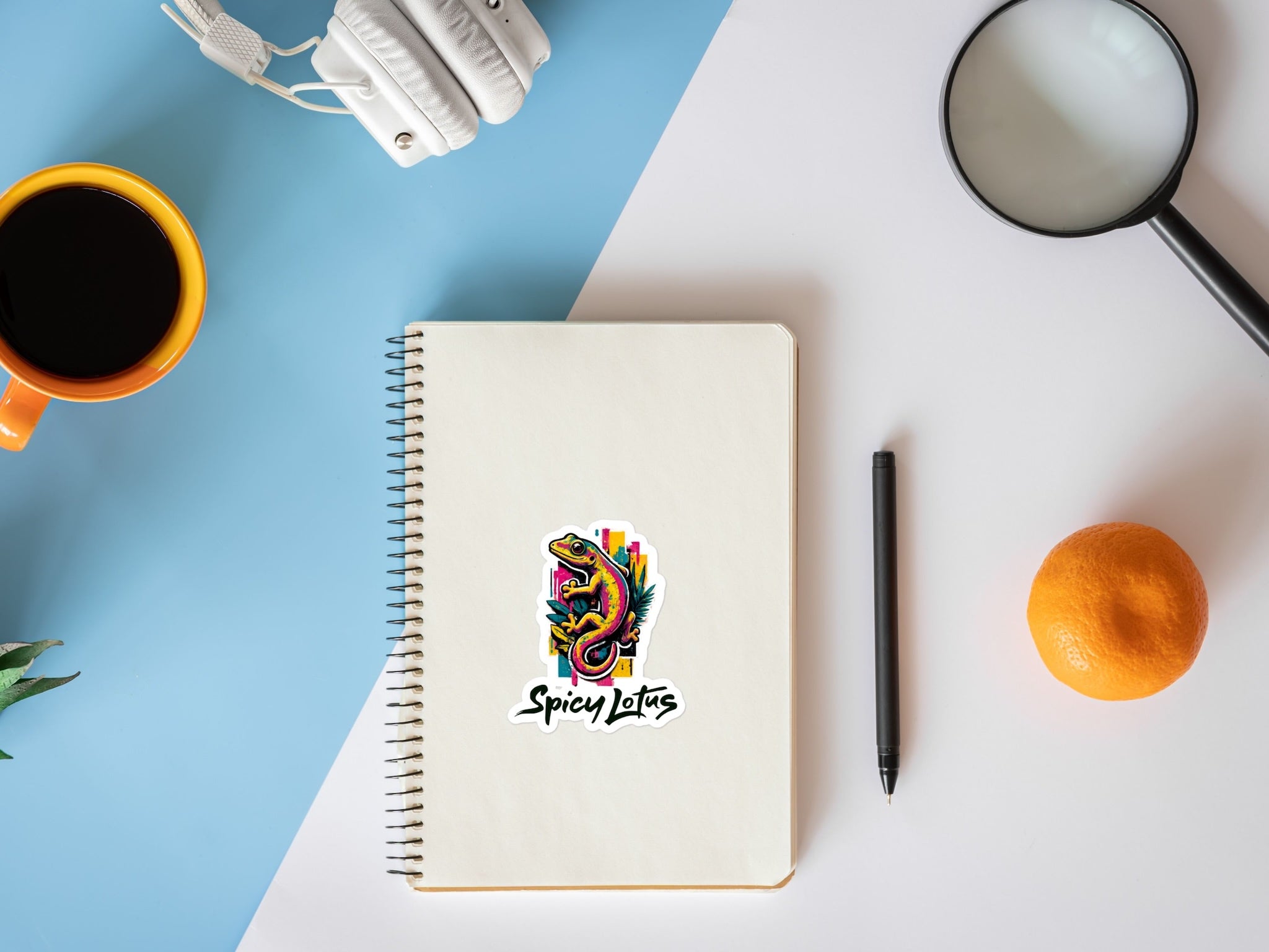Notebook with 'Spicy Lotus' logo with vibrant gecko graphic on a desk with coffee, pen, and orange.
