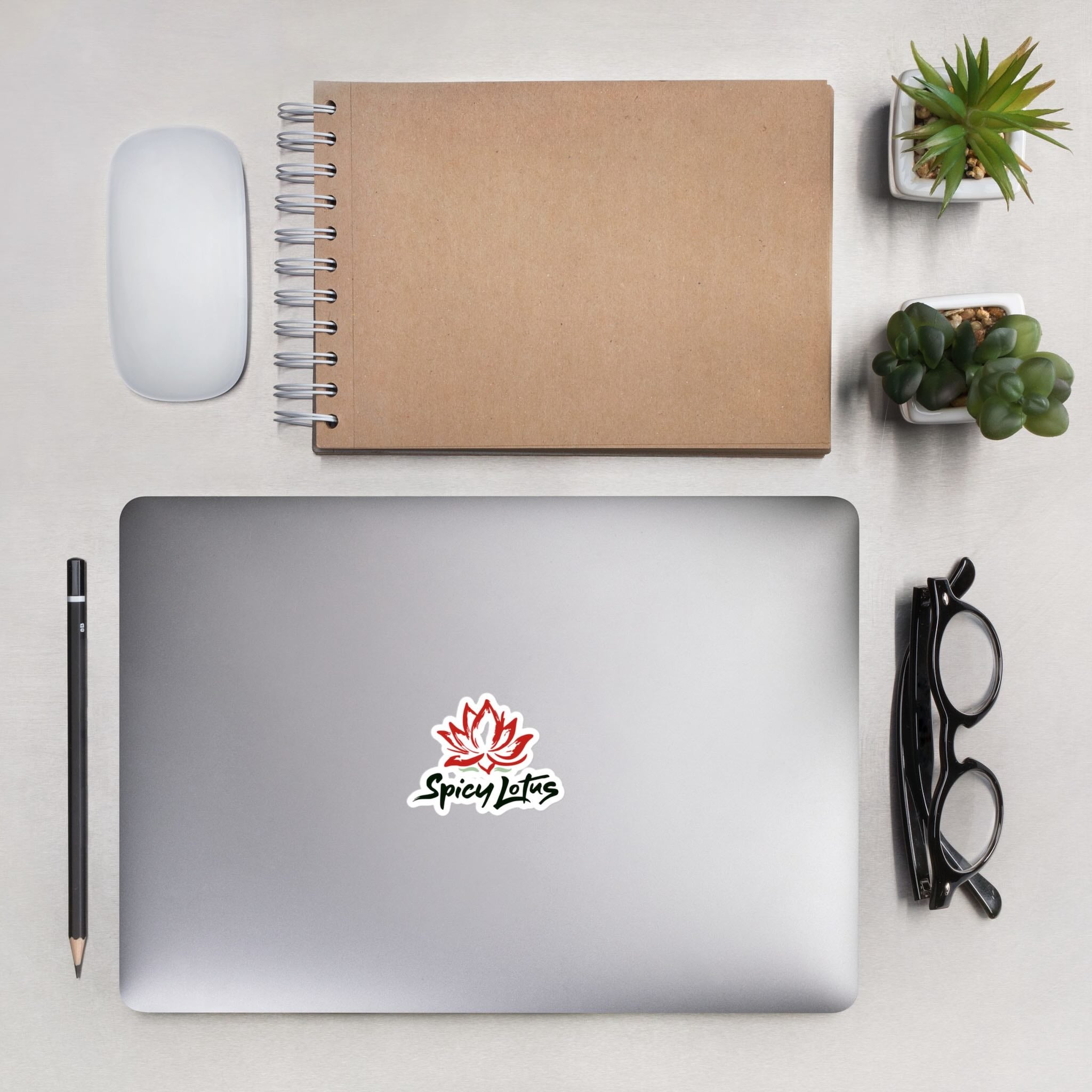 Laptop with Spicy Lotus logo sticker with brand name and red lotus flower on  a notebook,, next to  mouse, pencil, and glasses on white desk.