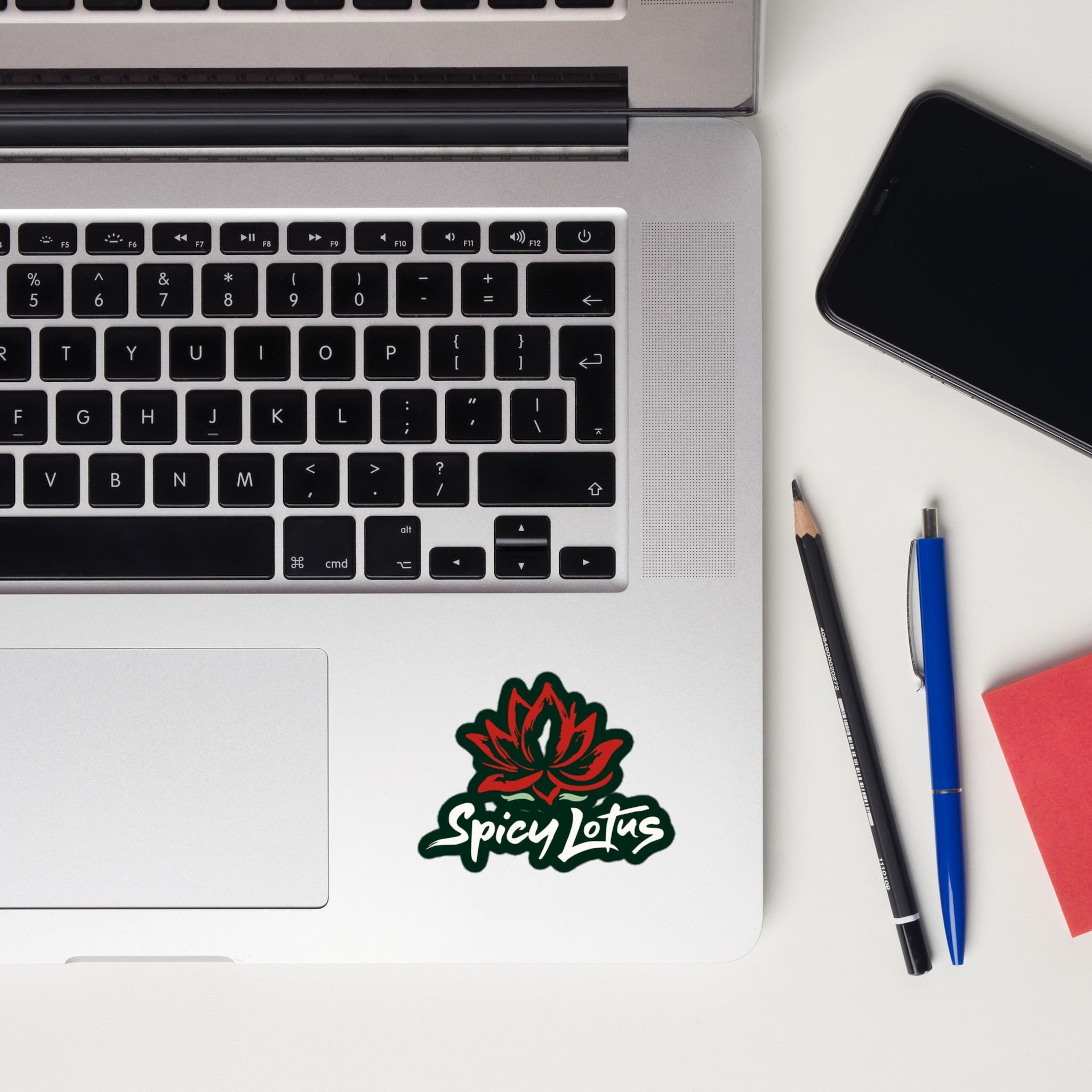 Laptop with Spicy Lotus sticker with lotus flower and brand name in white font on a notebook next to a phone, pencil, and pen.