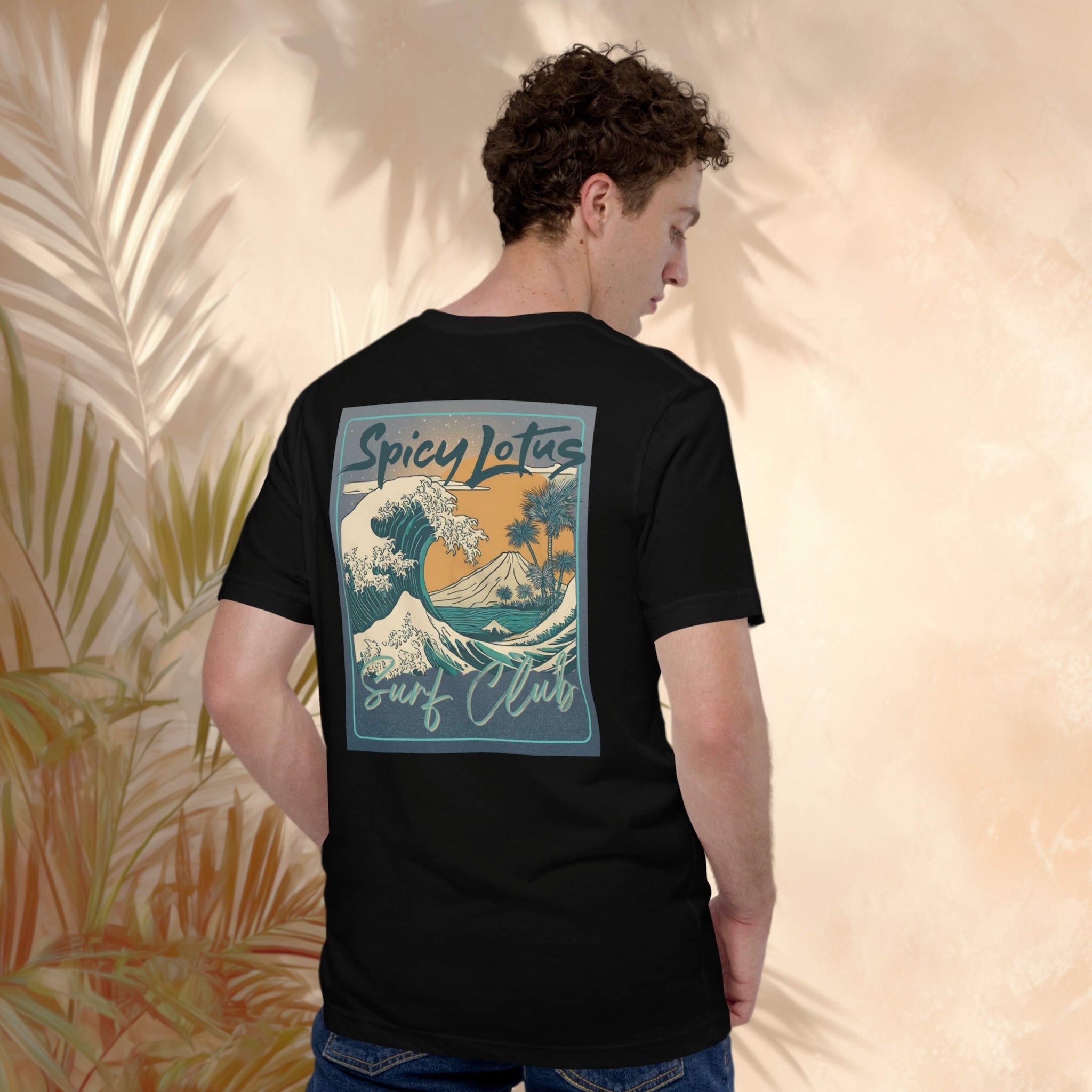 Person wearing a black t-shirt with a colorful Spicy Lotus Surf Club Bali volcano graphic on the back, standing against a beige wall with palm leaves.