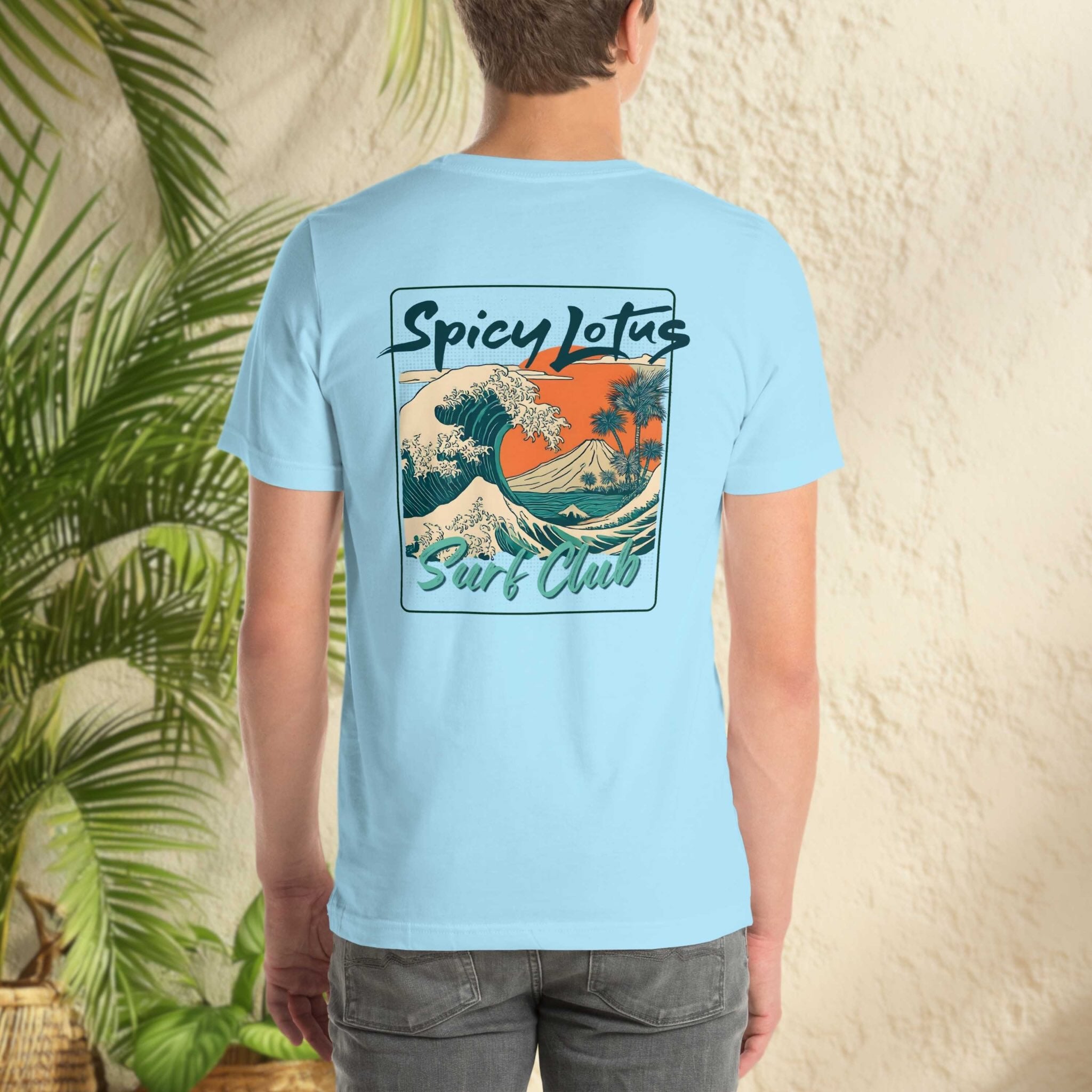 Person wearing a light blue t-shirt with 'Spicy Lotus Surf Club' design, standing against a textured wall with greenery.