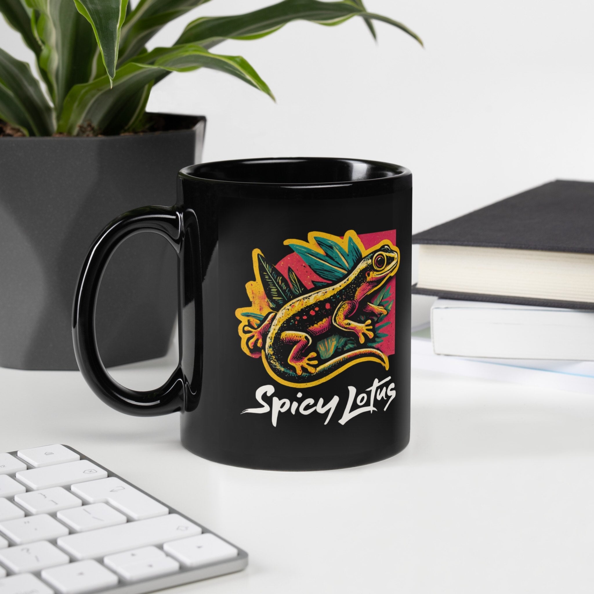 Black mug with colorful gecko design and 'Spicy Lotus' text on a desk with a plant and books in the background.