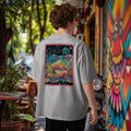 Man wearing a gray oversized t-shirt with a colorful graphic of vietamese summer rolls on the back, standing in an lush outdoor setting with a graffiti mural and tables.