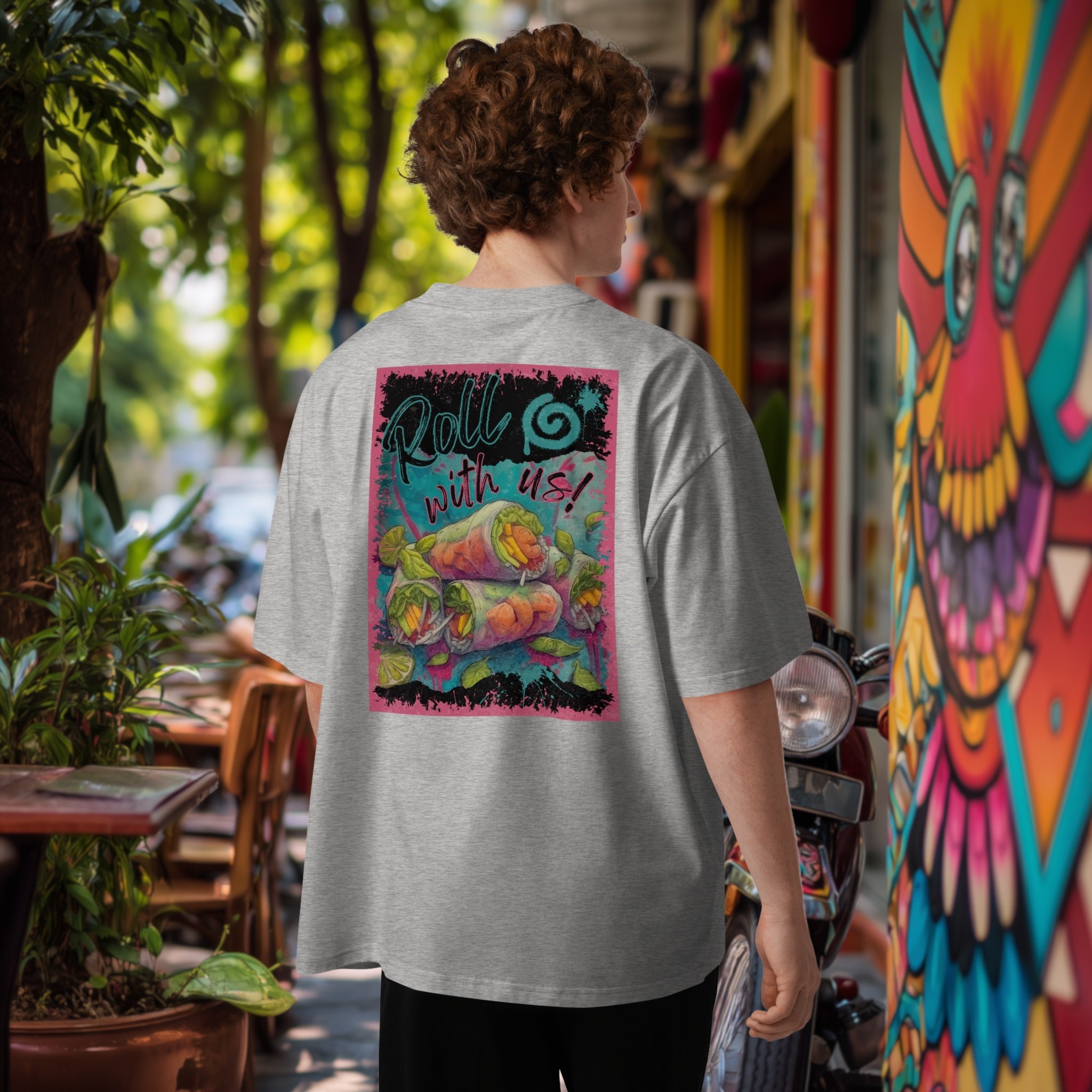 Man wearing a gray oversized t-shirt with a colorful graphic of vietamese summer rolls on the back, standing in an lush outdoor setting with a graffiti mural and tables.