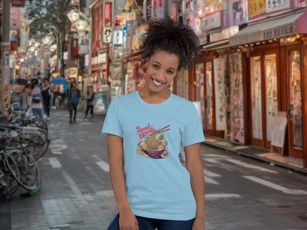 Person wearing a light blue t-shirt with a colorful graphic design of Ramen with 'Send Noods' in an urban street setting.