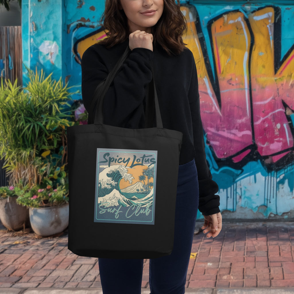 Woman holding a black tote bag with 'Spicy Lotus Surf Club' design in front of a colorful mural.