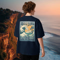 Male model wearing a navy blue t-shirt with 'Spicy Lotus Surf Club' design, standing on a cliff overlooking the ocean.