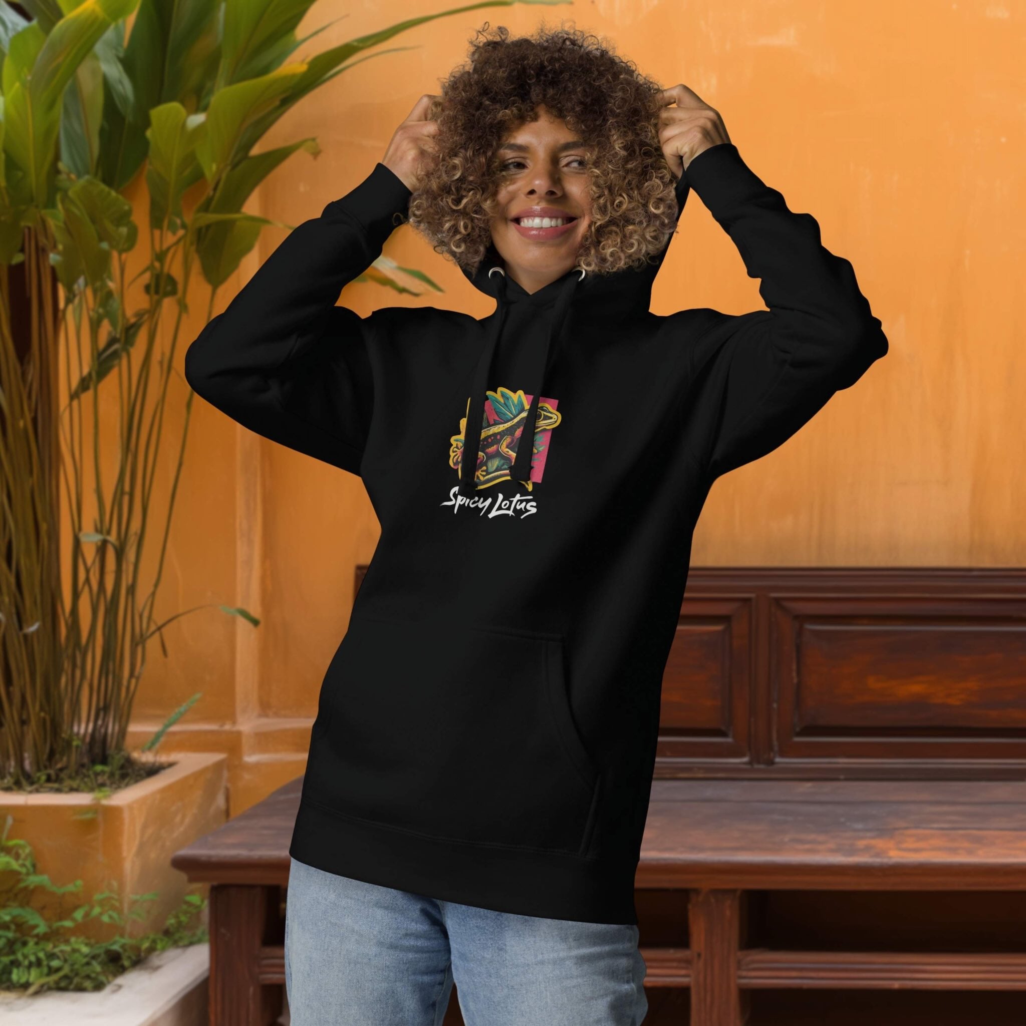 Person wearing a black hoodie with colorful design of geckos and tbrand name 'Spicy Lotus', standing against an orange wall with a wooden bench and plant.
