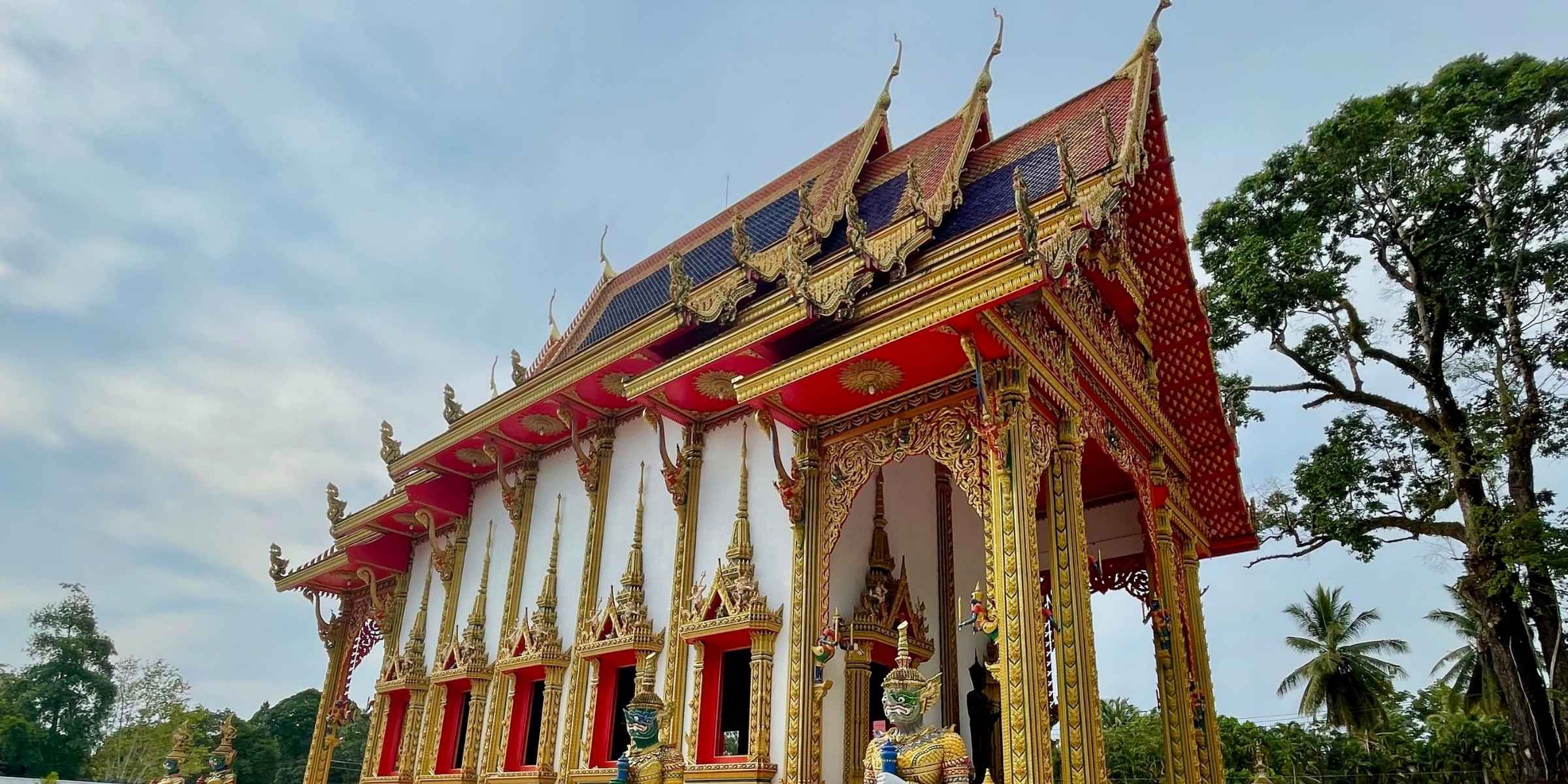 White Thai temple with gold and red accents on roof with lush greenery around