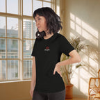 Person wearing a black t-shirt with a logo in a bright room with large windows and plants.