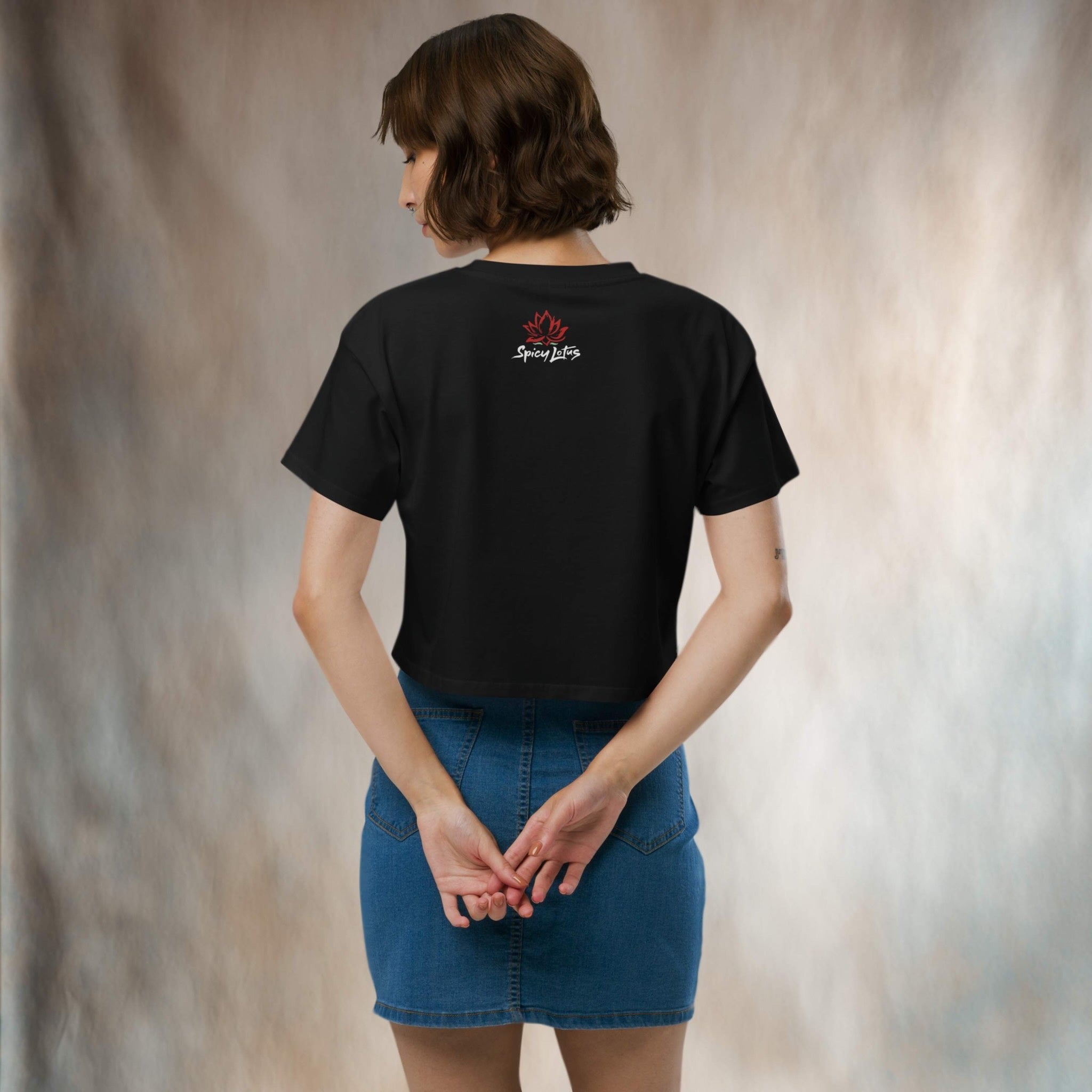 Person wearing a black crop tee with a logo on the back, standing in front of a neutral backdrop.